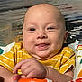 baby, child, smiling, tongue_out, yellow_shirt, toy, hand, face, dimples, bald_head, colorful, fabric, patterned, infant, cute, playful, indoors, closeup, person, happy