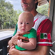 Jacob Ap is registered to the contest to win money with this photo: baby, man, holding, green_clothing, red_shirt, mexican_flag, outdoor, raindrops, balcony, hand, portrait, face, expression, infant, person, nature, wooden_railing, short_sleeves, daylight, skin