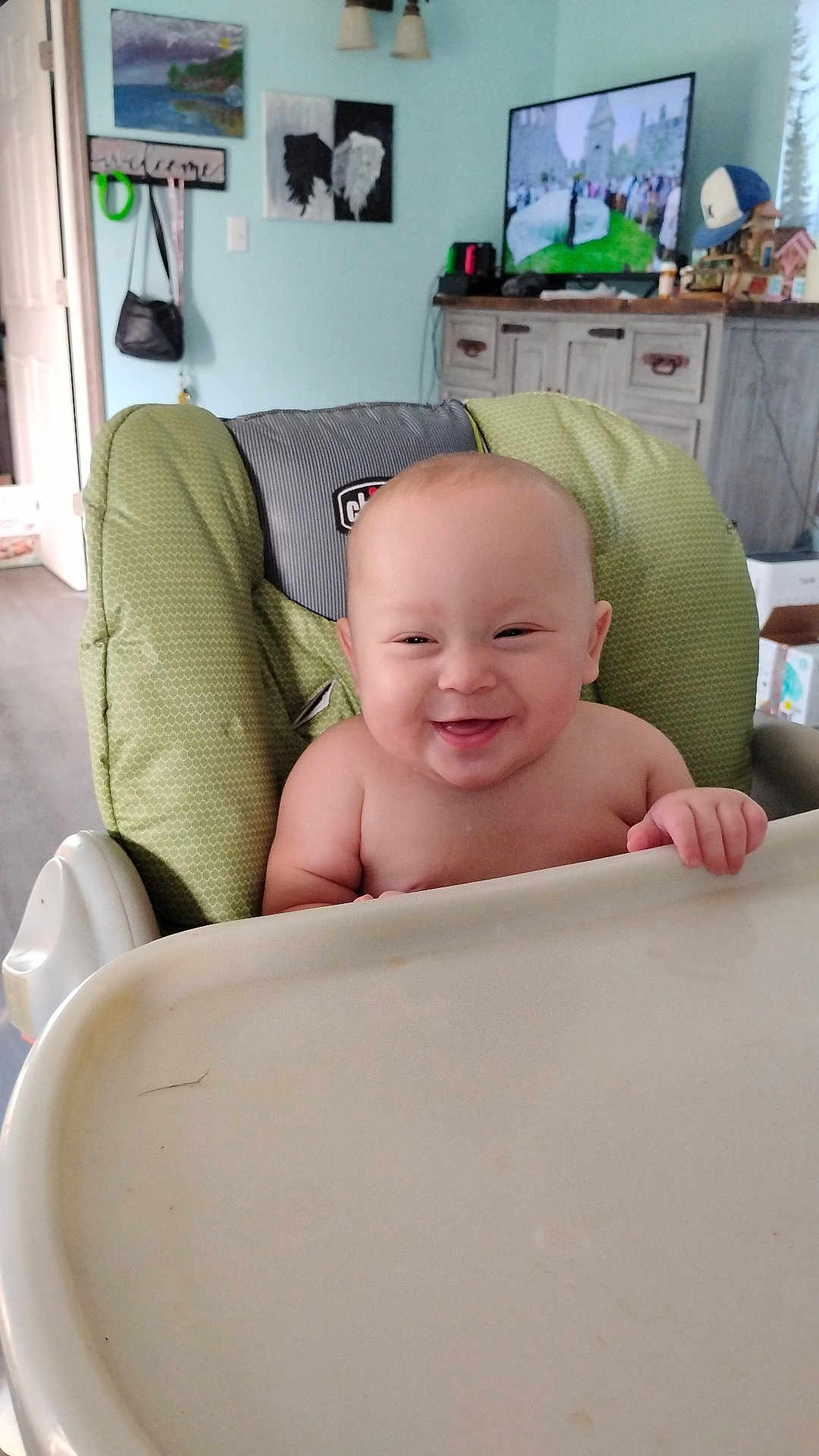 Jacob Ap is registered to the contest to win money with this photo: baby, high_chair, smiling, happy, indoor, child, face, furniture, tray, living_room, television, painting, decor, hand, skin, person, seat, expression, head, room