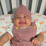 Gabriela is registered to the contest to win money with this photo: baby, smiling, knitted_hat, pink_outfit, butterfly_pattern, cushion, crib, stripes, child, happy, infant, cute, portrait, indoors, soft_lighting, apparel, button, hand, face, skin