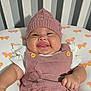 baby, smiling, knitted_hat, pink_outfit, butterfly_pattern, cushion, crib, stripes, child, happy, infant, cute, portrait, indoors, soft_lighting, apparel, button, hand, face, skin