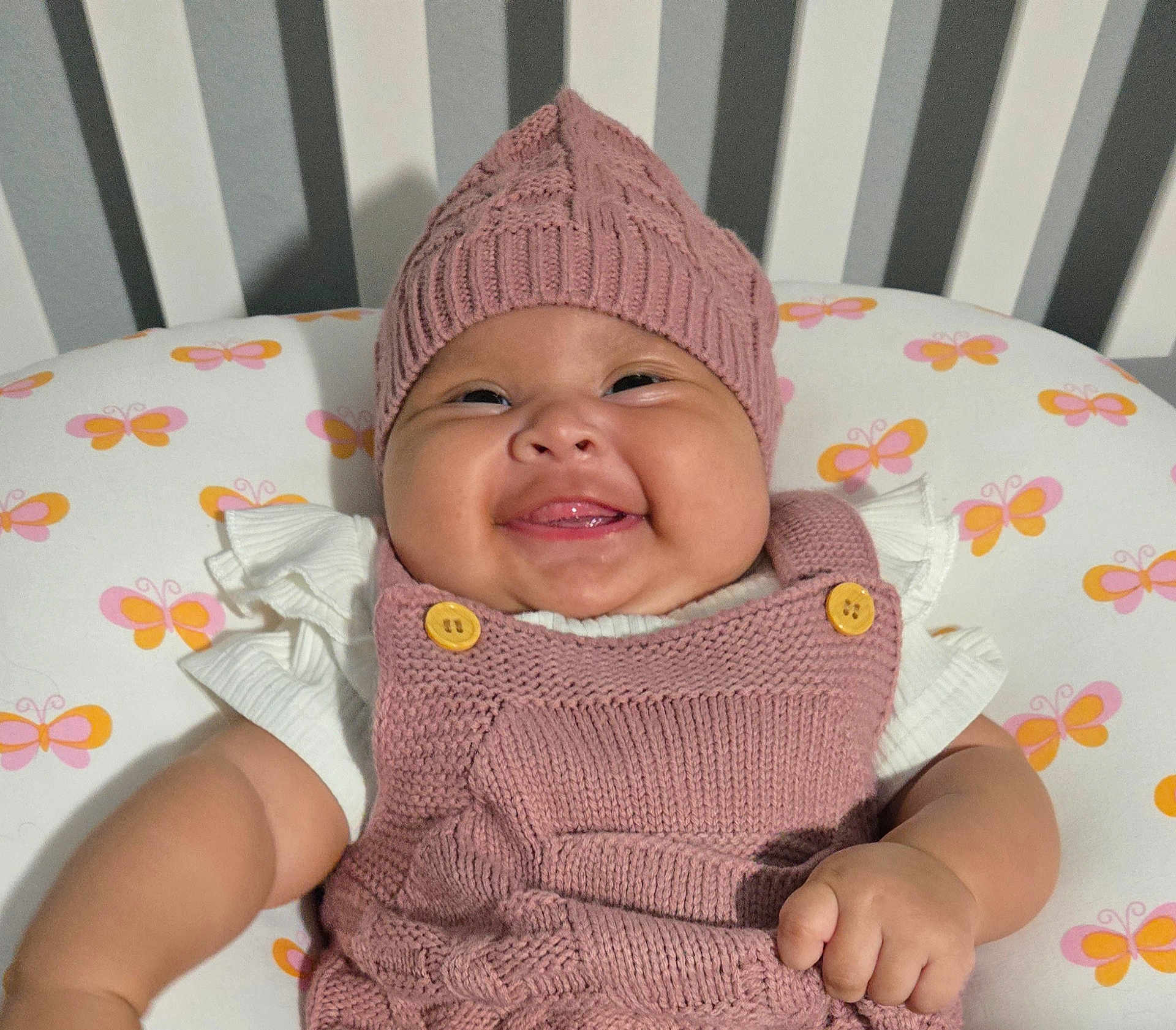 Gabriela is registered to the contest to win money with this photo: baby, smiling, knitted_hat, pink_outfit, butterfly_pattern, cushion, crib, stripes, child, happy, infant, cute, portrait, indoors, soft_lighting, apparel, button, hand, face, skin