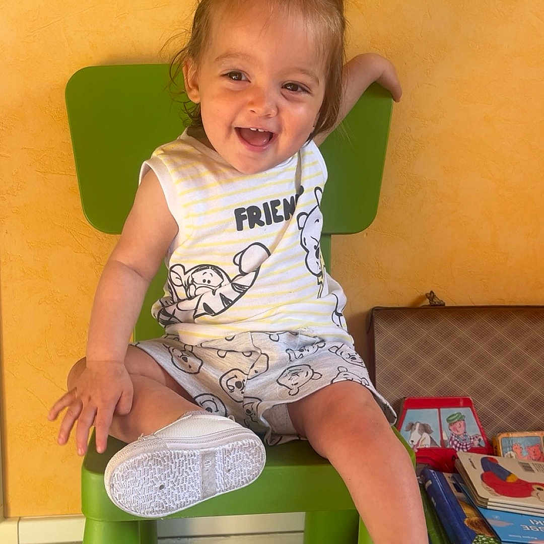 Kelly participe au concours pour gagner de l'argent avec cette photo : books, child, floor, footwear, furniture, green_chair, hair, happy, indoor, person, playful, room, shorts, sitting, sleeveless_shirt, smiling, stack, toddler, white_shoes, yellow_wall