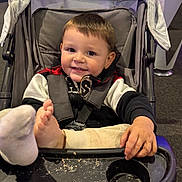 Malakai is registered to the contest to win money with this photo: toddler, child, stroller, smile, foot, clothing, seat, tray, crumbs, indoor, playful, baby, person, young_child, casual, happy, black_jacket, beige_pants, seatbelt, hand