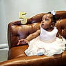 baby, child, girl, dress, white_dress, pearl_belt, couch, leather_couch, brown_couch, number_five, golden_number, birthday, celebration, sitting, reaching, curious, earrings, hair_bow, indoor, portrait