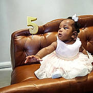 Farida is registered to the contest to win money with this photo: baby, child, dress, white_dress, couch, leather_couch, birthday, number_five, candle, pearl_belt, hair_bow, curious, sitting, indoor, celebration, portrait, cute, person, young_child, furniture