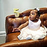 baby, child, dress, white_dress, couch, leather_couch, birthday, number_five, candle, pearl_belt, hair_bow, curious, sitting, indoor, celebration, portrait, cute, person, young_child, furniture