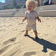 Zayn a rejoint le concours — aidez-le/la à gagner de superbes lots ! toddler, child, blonde_hair, curly_hair, smiling, sand, beach, sunny_day, shadow, nike_clothing, shorts, tshirt, footwear, urban_background, people, outdoor, daylight, happy, standing, playful