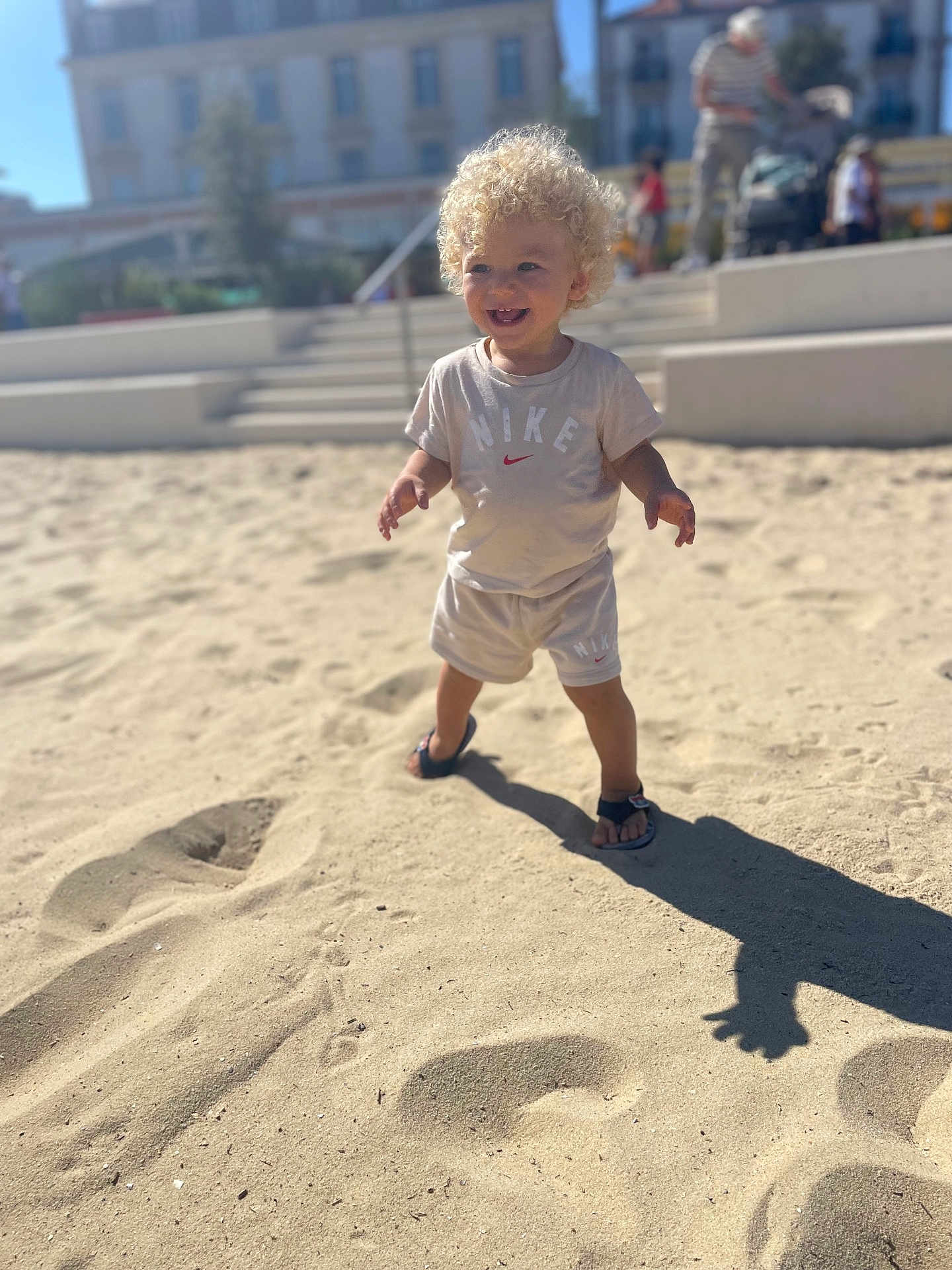 Zayn a rejoint le concours — aidez-le/la à gagner de superbes lots ! toddler, child, blonde_hair, curly_hair, smiling, sand, beach, sunny_day, shadow, nike_clothing, shorts, tshirt, footwear, urban_background, people, outdoor, daylight, happy, standing, playful
