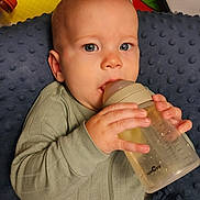 Luka is registered to the contest to win money with this photo: baby, infant, child, bottle, feeding, formula, sitting, lying_down, wide_eyed, face, hands, gray_outfit, blanket, nursery, toys, colorful, indoors, portrait, close_up, happy