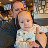 Luka joined the competition — help win amazing prizes! baby, infant, adult, father, portrait, blue_eyes, hoodie, onesie, baby_clothing, holding, indoor, restaurant, bar, menu_board, framed_art, warm_lighting, seated, close_up, family, photograph