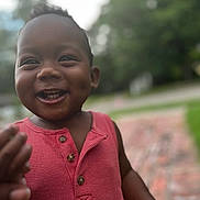 Jireh is registered to the contest to win money with this photo: child, toddler, smiling, happy, face, dimples, red_shirt, sleeveless, outdoor, daylight, portrait, close_up, person, skin, emotion, joy, cute, young_child, buttoned_shirt, blurred_background