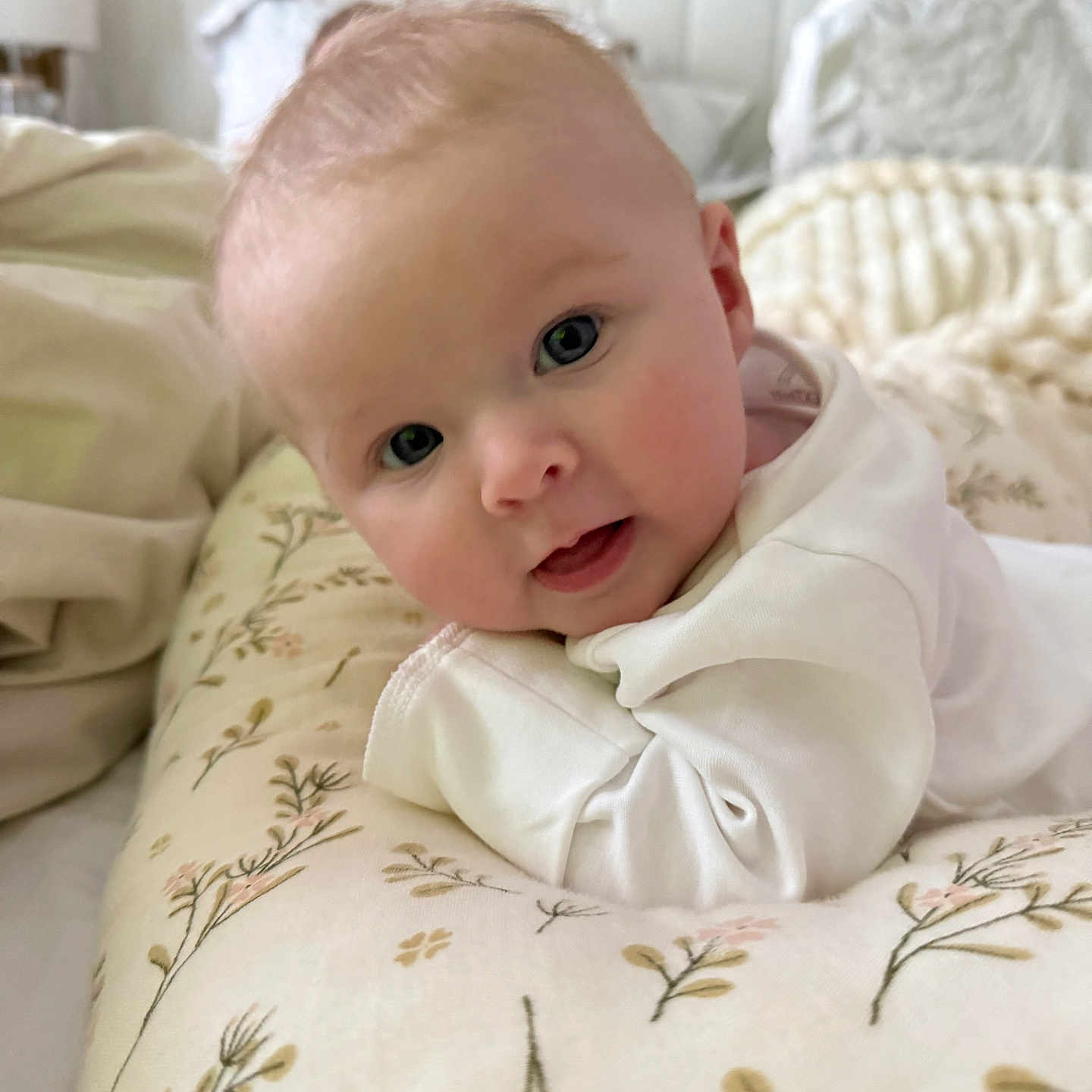 Eliza joined the competition — help win amazing prizes! baby, bedroom, blanket, child, cozy, curious, cushion, cute, eyes, face, floral_pattern, head_support, indoors, infant, lying_down, person, portrait, smile, soft_lighting, white_clothing