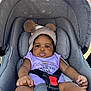 baby, infant, car_seat, hat, pom_poms, purple_clothing, straps, outdoor, child, seatbelt, cute, face, person, sitting, expression, headwear, safety, young_child, portrait, daylight