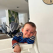 Alex a rejoint le concours — aidez-le/la à gagner de superbes lots ! baby, high_chair, striped_clothing, smiling, indoor, stroller, wall_clock, wood_floor, hand_on_face, cheeky, young_child, playful, modern_home, leafy_wallpaper, furniture, cute, face, person, relaxed, casual