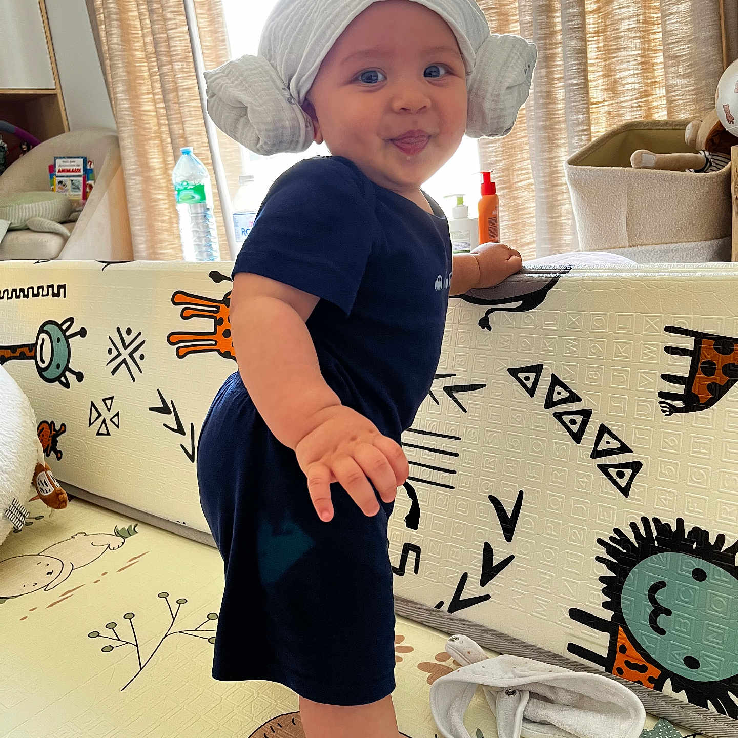 Léo participe au concours pour gagner de l'argent avec cette photo : animal_print, baby, baby_item, bottle, child, curtains, expression, face, foot, furniture, hand, head_wrap, indoor, navy_clothing, playmat, room, smiling, standing, toddler, toy