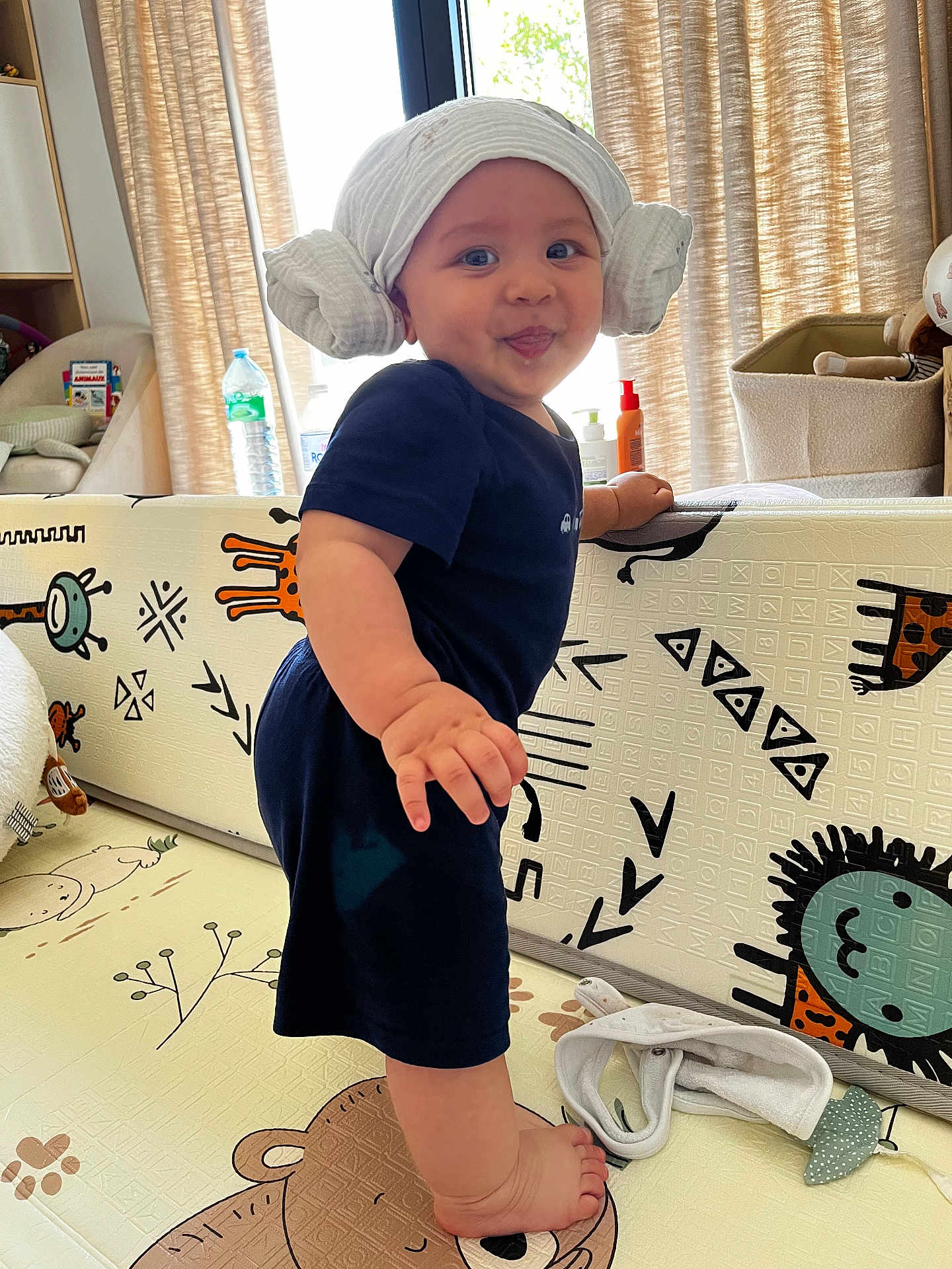 Léo participe au concours pour gagner de l'argent avec cette photo : baby, toddler, standing, head_wrap, navy_clothing, playmat, animal_print, indoor, smiling, child, toy, curtains, bottle, furniture, foot, hand, face, room, baby_item, expression