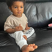 Elias joined the competition — help win amazing prizes! toddler, child, curly_hair, beige_top, white_pants, indoor, leather_couch, barefoot, serious_expression, sitting, young_child, person, portrait, casual_clothing, home, furniture, relaxed, cute, small_toes, focused