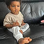 toddler, child, curly_hair, beige_top, white_pants, indoor, leather_couch, barefoot, serious_expression, sitting, young_child, person, portrait, casual_clothing, home, furniture, relaxed, cute, small_toes, focused