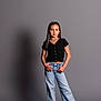 child, girl, standing, black_top, blue_jeans, sneakers, casual_clothing, studio, gray_background, portrait, smile, long_hair, fashion, hands_on_hips, full_body, shadow, young, confident, pose, indoor