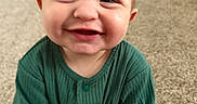 Riot is registered to the contest to win money with this photo: baby, child, smiling, green_clothing, ribbed_fabric, carpet, indoor, toy, happy, face, hair, spiky_hair, person, infant, cute, sitting, floor, home, furniture, car_seat