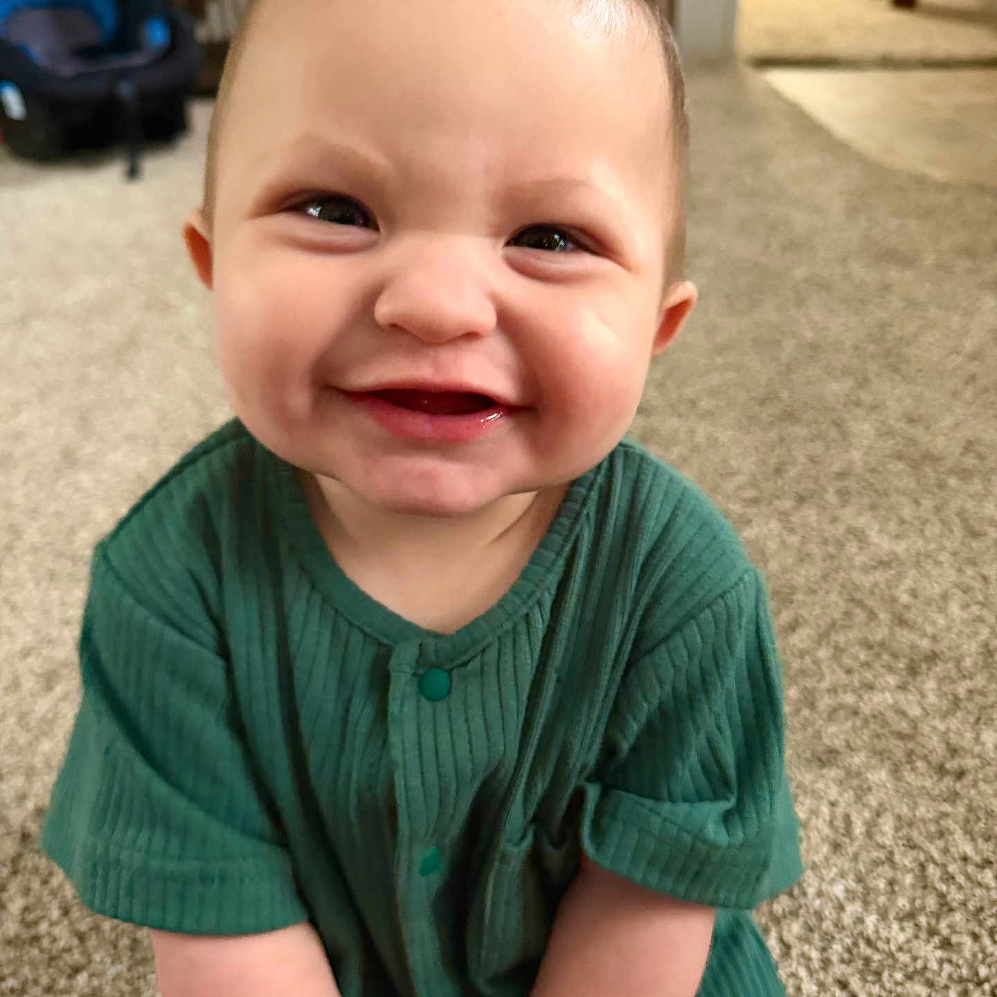 Riot is registered to the contest to win money with this photo: baby, child, smiling, green_clothing, ribbed_fabric, carpet, indoor, toy, happy, face, hair, spiky_hair, person, infant, cute, sitting, floor, home, furniture, car_seat