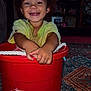 Jakarii is registered to the contest to win money with this photo: toddler, child, smiling, curly_hair, red_bucket, indoor, carpet, playful, happy, face, person, sitting, toy, television, animation, room, furniture, home, casual_clothing, hands