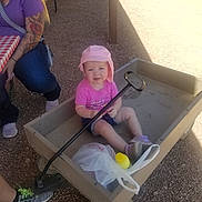 Harper is registered to the contest to win money with this photo: toddler, child, wagon, pink_hat, pink_shirt, shorts, shoes, adult, tattoos, purple_shirt, picnic_table, checkered_tablecloth, outdoor, gravel, sunlight, shadow, plastic_bag, yellow_bottle, handle, seated