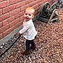 Harper is registered to the contest to win money with this photo: toddler, child, red_hair, smiling, outdoor, pebbles, brick_wall, hose, hose_reel, boots, white_shirt, dark_pants, curious, standing, playful, garden, side_view, daylight, person, young_child