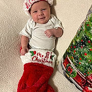 Emma joined the competition — help win amazing prizes! baby, christmas, stocking, santa_hat, white_onesie, holiday, smiling, face, infant, blanket, decor, festive, red, white, cozy, cute, indoors, pillow, christmas_tree, holiday_season