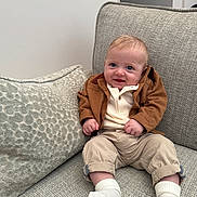 Charlie joined the competition — help win amazing prizes! baby, child, smile, blue_eyes, brown_jacket, beige_pants, white_socks, couch, pillow, indoor, infant, cute, portrait, seated, happy, cozy, homedecor, textured_fabric, person, small_hands