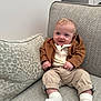 baby, child, smile, blue_eyes, brown_jacket, beige_pants, white_socks, couch, pillow, indoor, infant, cute, portrait, seated, happy, cozy, homedecor, textured_fabric, person, small_hands
