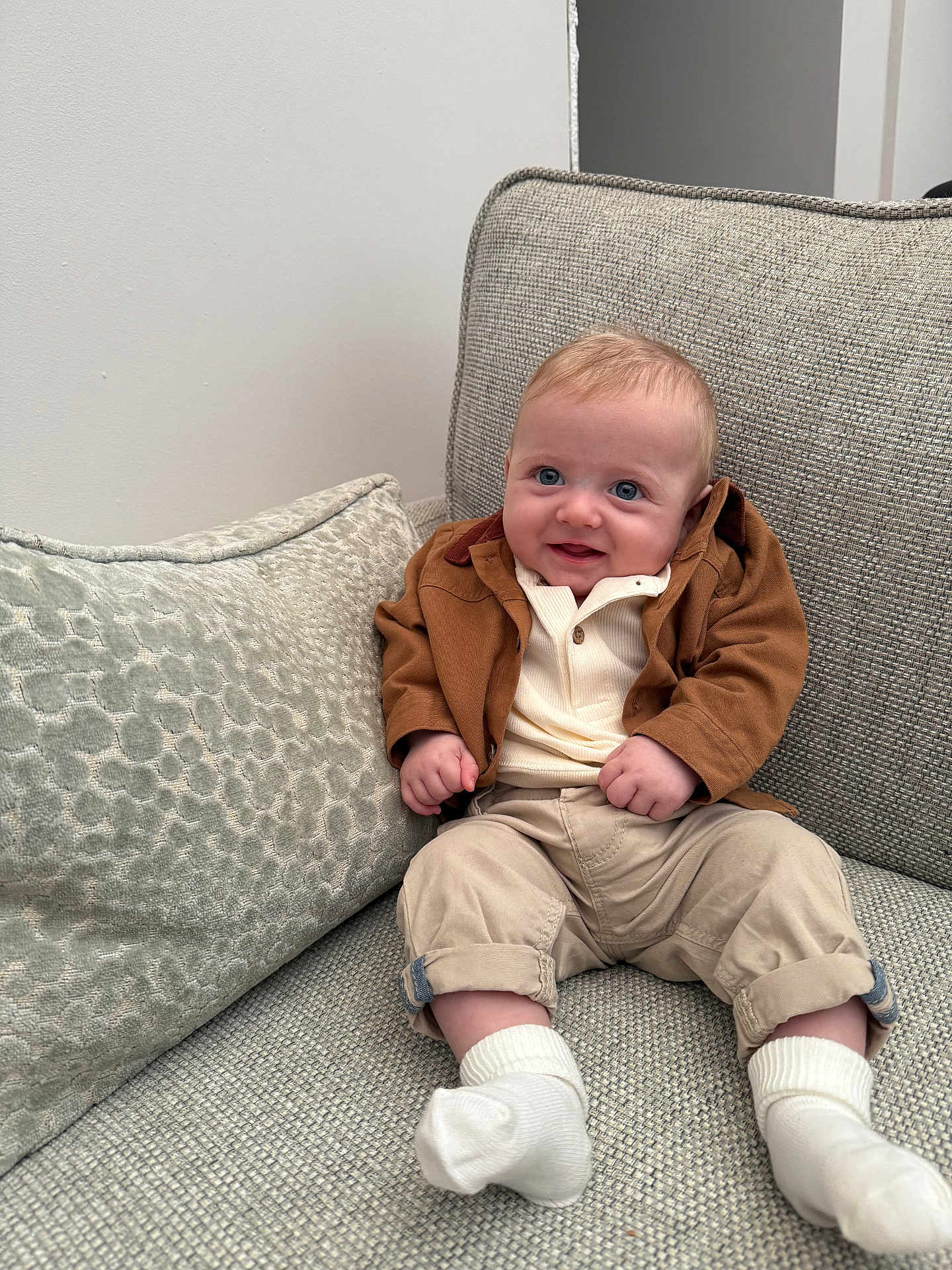 Charlie joined the competition — help win amazing prizes! baby, child, smile, blue_eyes, brown_jacket, beige_pants, white_socks, couch, pillow, indoor, infant, cute, portrait, seated, happy, cozy, homedecor, textured_fabric, person, small_hands