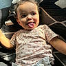 Soraya joined the competition — help win amazing prizes! baby, toddler, child, curly_hair, tongue_out, smiling, couch, pillow, patterned_fabric, indoor, natural_light, short_sleeve_shirt, diaper, playful, happy, skin, face, person, seated, cozy