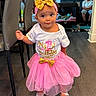 birthday, boy, celebration, child, couch, curly_hair, decor, family, flooring, gold_bow, headband, home, indoor, person, pink_tutu, playpen, shoes, smiling, toddler, white_shirt