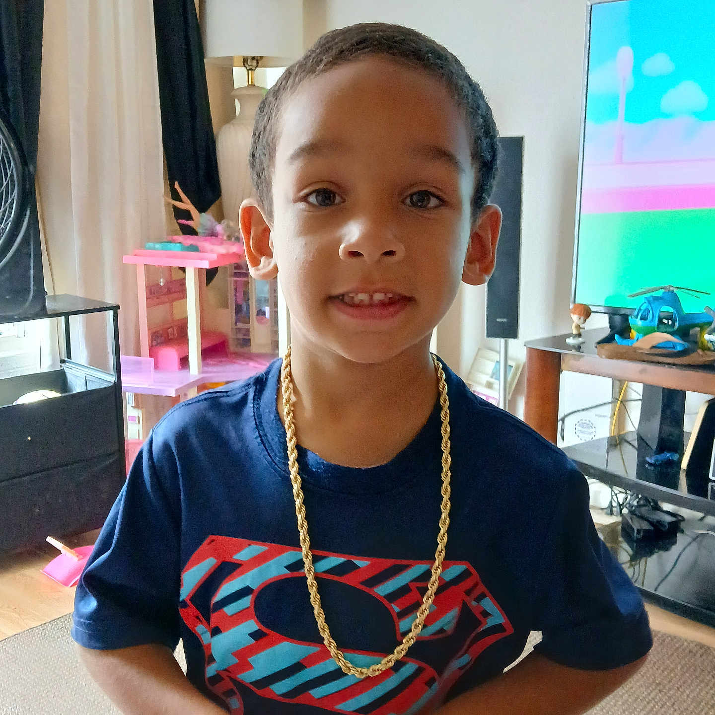 Shyden is registered to the contest to win money with this photo: child, boy, superman_shirt, gold_chain, smile, indoor, living_room, toy, television, furniture, carpet, lamp, curtain, table, speaker, person, portrait, casual, home, happy