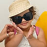 Khloe joined the competition — help win amazing prizes! child, sunglasses, hat, visor, smile, hands, yellow_chair, white_shirt, curly_hair, indoor, playful, person, portrait, accessory, fashion, cute, happy, casual, young, fun
