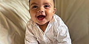 Ronald is registered to the contest to win money with this photo: baby, infant, child, smiling, happy, sitting, white_shirt, barefoot, indoors, couch, soft_furniture, curly_hair, cute, portrait, playful, young_child, skin, hand, foot, cozy