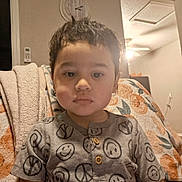 Luis is registered to the contest to win money with this photo: toddler, child, boy, face, expressionless, shirt, smiley_faces, peace_signs, blanket, chair, indoor, living_room, clock, light, wall, person, hair, sitting, casual_clothing, portrait