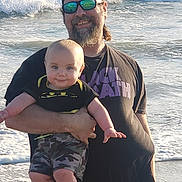 Ashton is registered to the contest to win money with this photo: beach, baby, man, ocean, waves, sunglasses, headband, camouflage, smile, holding, water, outdoor, child, casual_clothing, sunlight, person, happy, father, infant, summer