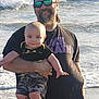 beach, baby, man, ocean, waves, sunglasses, headband, camouflage, smile, holding, water, outdoor, child, casual_clothing, sunlight, person, happy, father, infant, summer