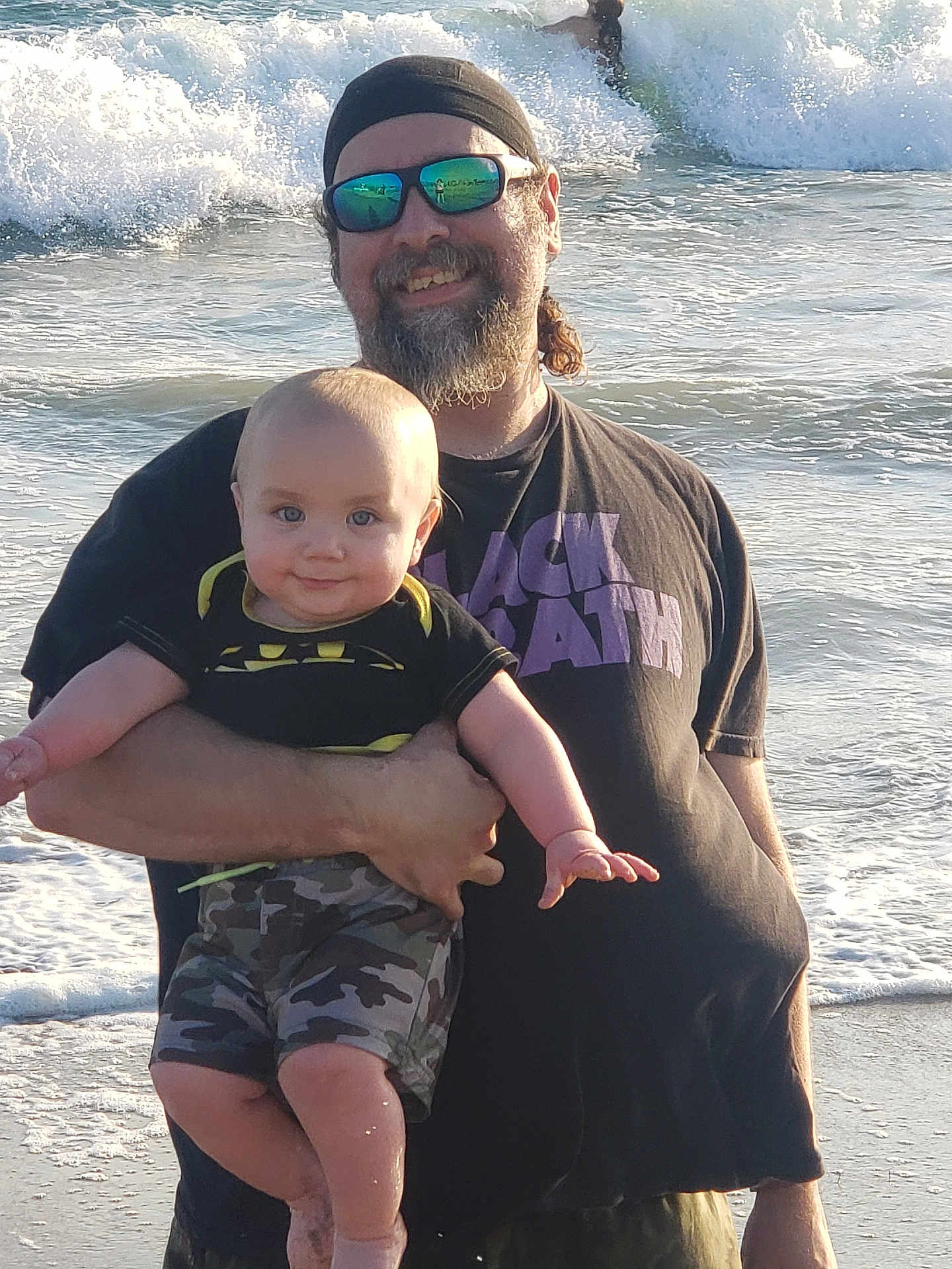 Ashton is registered to the contest to win money with this photo: beach, baby, man, ocean, waves, sunglasses, headband, camouflage, smile, holding, water, outdoor, child, casual_clothing, sunlight, person, happy, father, infant, summer