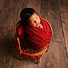 newborn, baby, sleeping, infant, portrait, red_blanket, swaddled, wicker_chair, basket, wooden_floor, indoors, peaceful, cozy, closed_eyes, black_hair, head, hands, newborn_photography, warm_lighting, adorable
