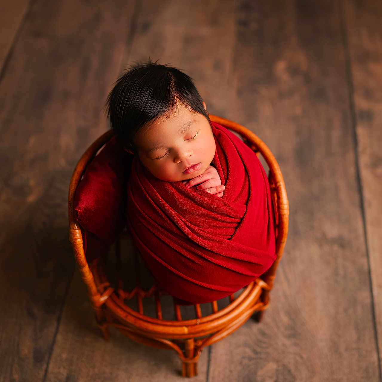 Niall is registered to the contest to win money with this photo: baby, newborn, sleeping, infant, portrait, red_blanket, swaddled, basket, wicker, wooden_floor, indoor, overhead_view, peaceful, closed_eyes, black_hair, cozy, warm_lighting, hand, seated, photography