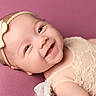 baby, bow, child, closeup, clothing, cute, face, happy, headband, indoors, infant, lace, lying_down, newborn, person, pink_background, portrait, skin, smiling, soft_background