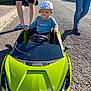Milo a rejoint le concours — aidez-le/la à gagner de superbes lots ! toddler, toy_car, outdoor, street, adult_legs, bucket_hat, pacifier, sunny, sidewalk, vehicle, green, jeans, shorts, footwear, asphalt, child, daylight, residential_area, people, playing