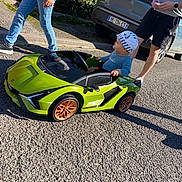 Milo participe au concours pour gagner de l'argent avec cette photo : toddler, toy_car, green_car, hat, pacifier, sidewalk, street, adult, shadow, sunlight, casual_clothing, jeans, shorts, sneakers, nature, hedge, vehicle, license_plate, parking, outdoor