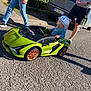 toddler, toy_car, green_car, hat, pacifier, sidewalk, street, adult, shadow, sunlight, casual_clothing, jeans, shorts, sneakers, nature, hedge, vehicle, license_plate, parking, outdoor