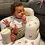 baby, child, curly_hair, inflatable_chair, teddy_bear_pattern, snack, bite, funny_face, living_room, couch, carpet, white_clothing, green_pants, bare_feet, indoor, cute, infant, seated, playful, cozy