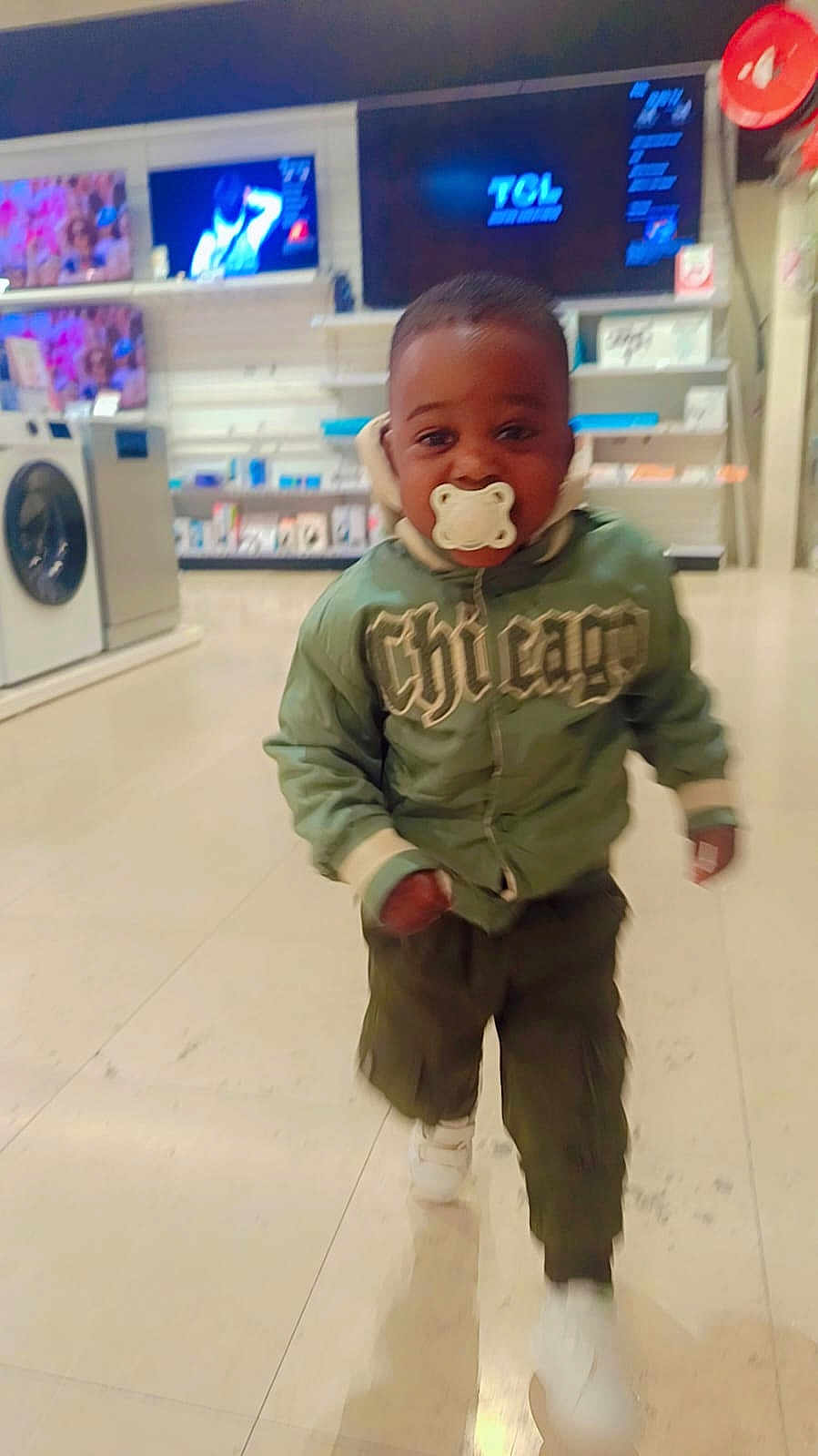 Ychane participe au concours pour gagner de l'argent avec cette photo : toddler, child, pacifier, green_jacket, walking, indoor, store, television, appliances, floor, white_shoes, pants, face, person, motion_blur, shopping, electronics, cute, young, hoodie